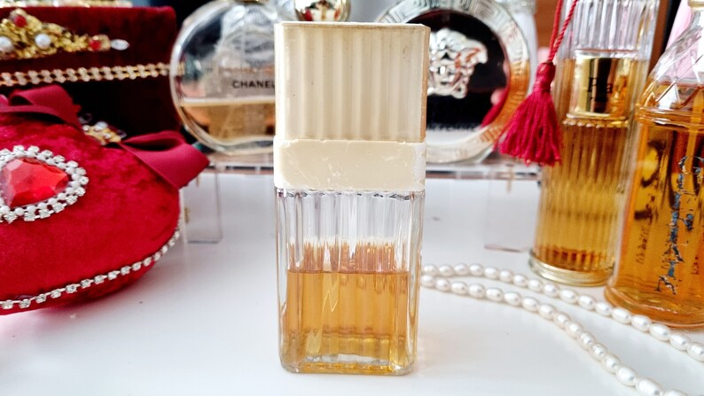 May include: A clear glass bottle of perfume with a gold cap and a white label. The bottle is filled with a light amber liquid. The bottle is sitting on a white surface.