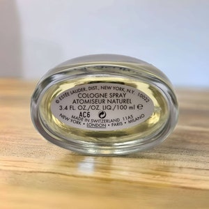 Może przedstawiać: Przezroczysta butelka szklana z złotą zakrętką i białą etykietą. Etykieta zawiera napis "COLOGNE SPRAY ATOMISEUR NATUREL 3.4 FL. OZ./OZ. LIQ./100 ml e AC6 MADE IN SWITZERLAND 11A5 NEW YORK LONDON PARIS MILANO"