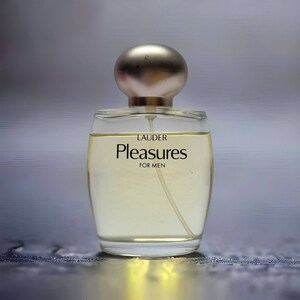 Może przedstawiać: Przezroczysta szklana butelka perfum z srebrną zakrętką. Na butelce widnieją napisy "LAUDER" i "Pleasures", a także słowa "FOR MEN".
