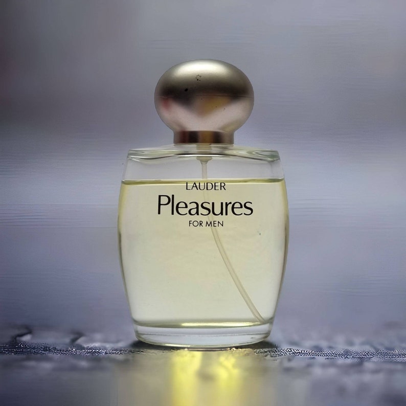 Może przedstawiać: Przezroczysta szklana butelka perfum z srebrną zakrętką. Na butelce widnieją napisy "LAUDER" i "Pleasures", a także słowa "FOR MEN".
