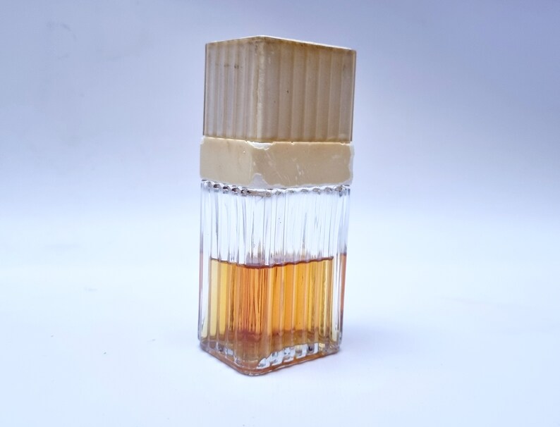 May include: A clear glass bottle with a textured surface and a beige plastic cap. The bottle is filled with a light brown liquid.