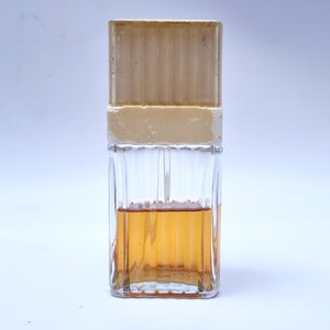 May include: A clear glass bottle with a beige cap and a brown liquid inside. The bottle has a textured design.
