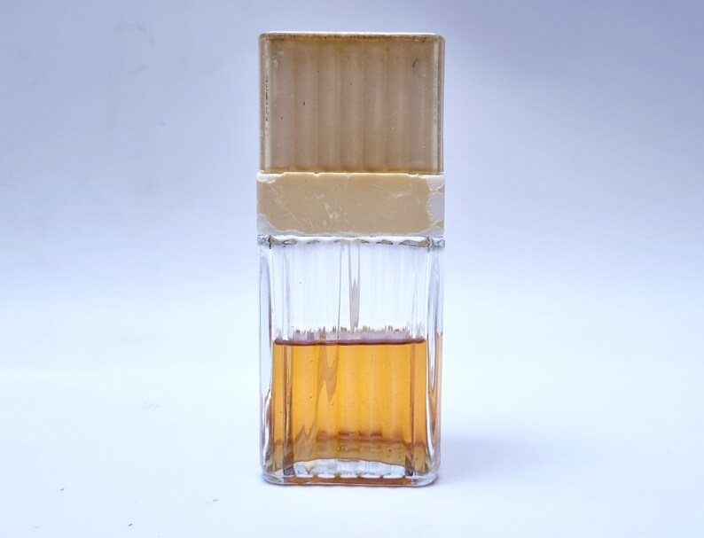May include: A clear glass bottle with a beige cap and a brown liquid inside. The bottle has a textured design.