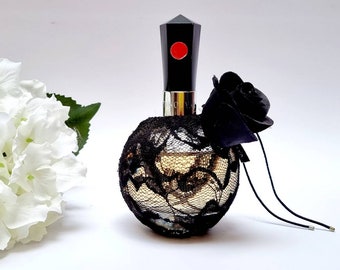 May include: A black bottle of perfume with a black lace design and a black rose on the top. The bottle is sitting on a white surface.