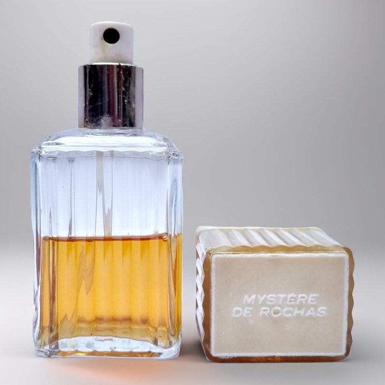 May include: A clear glass bottle of perfume with a silver cap and a white spray nozzle. The bottle is filled with a light amber liquid. The bottle is sitting on a white surface. The bottle is labeled "Mystere de Rochas".