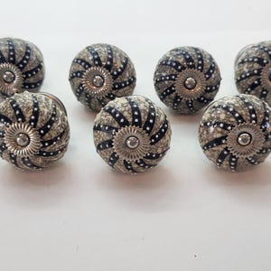 May include: A set of nine round cabinet knobs featuring a textured beige and grey crackle finish. Each knob has black radial stripes accented with small white dots and a central silver-toned metal circular plate with a ridged edge design.