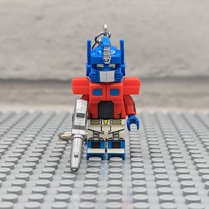 May include: A red, blue, and silver Lego minifigure of Optimus Prime, a robot character from the Transformers franchise. The minifigure is holding a silver gun and has a key chain attached.