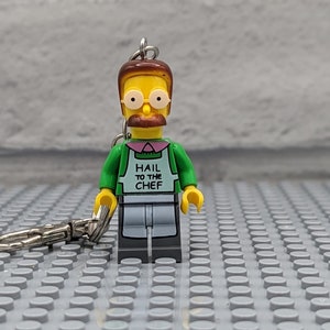 May include: A yellow Lego minifigure of a chef wearing a green shirt and an apron with the text "Hail to the Chef". The minifigure has brown hair, a mustache, and glasses. The minifigure is attached to a silver keychain.