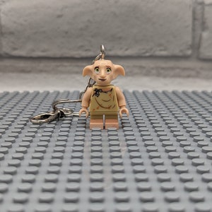 May include: A light brown Lego minifigure of Dobby the house-elf from the Harry Potter series. The minifigure is wearing a brown shirt and has a silver keychain attached to its back.