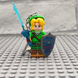 May include: A green and brown Lego minifigure of Link from the Legend of Zelda video game series. The minifigure is holding a blue sword and a shield with the Triforce symbol. The minifigure is attached to a keychain.