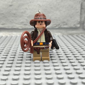 May include: A brown and tan Lego minifigure of an explorer wearing a brown hat and holding a whip and a gun. The minifigure is standing on a grey Lego baseplate.