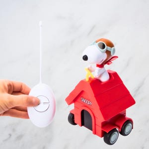 Puede incluir: Un juguete de coche con forma de caseta roja de Snoopy con mando a distancia. El juguete presenta a Snoopy con gafas y gorra de piloto. La caseta tiene la palabra "Snoopy" impresa. El mando a distancia es blanco.