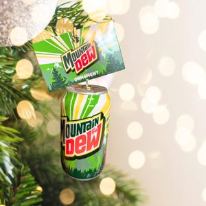 May include: A Mountain Dew soda can ornament with a matching tag. The can is green, white, and yellow with the Mountain Dew logo in red and green. The tag reads "Mountain Dew Ornament" with a forest scene.