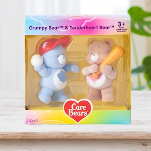 May include: A boxed set of Care Bears toys featuring Grumpy Bear and Tenderheart Bear. Grumpy Bear is blue and holding a baseball and wearing a red cap. Tenderheart Bear is brown and holding a baseball bat. The box reads "Care Bears".