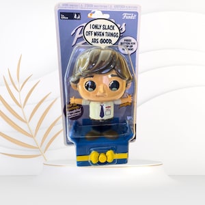 May include: A Funko Pop! figure of a man with brown hair wearing a blue shirt and a blue tie. The figure is sitting in a blue chair with a yellow button on the front. The packaging says "The Office" and "I only slack off when things are good."