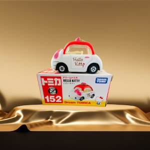 Könnte beinhalten: Ein weiß-rotes Hello Kitty Spielzeugauto mit dem Text "Hello Kitty" an der Seite, das auf einer Schachtel steht. Die Schachtel ist rot und weiß mit japanischem Text und der Nummer 152.