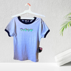 May include: Gray short-sleeved t-shirt with a black ringer neckline and a graphic of five cartoon characters wearing hats. The shirt has a Hello Kitty tag attached.