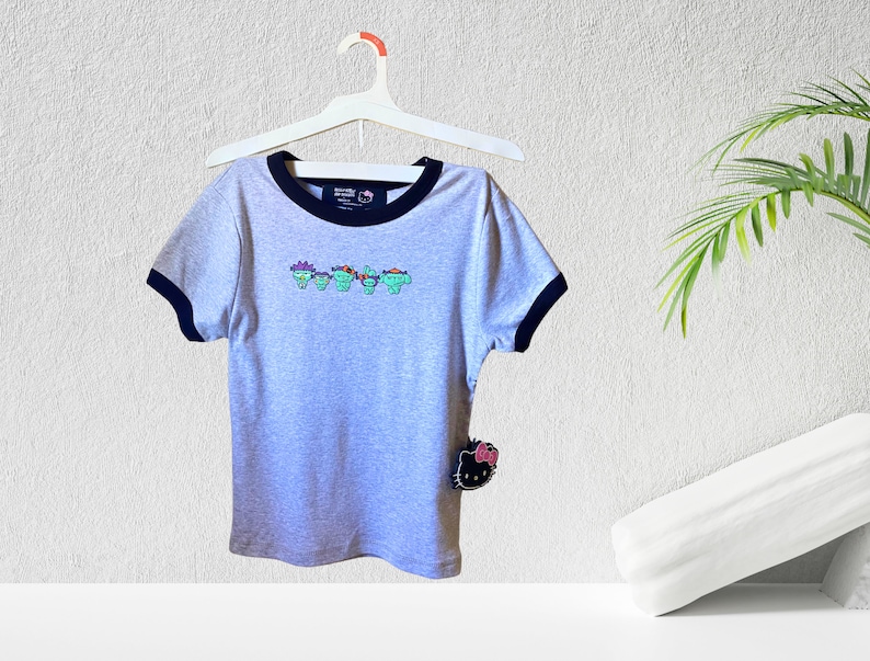 May include: Gray short-sleeved t-shirt with a black ringer neckline and a graphic of five cartoon characters wearing hats. The shirt has a Hello Kitty tag attached.