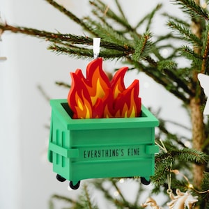 May include: A green dumpster Christmas ornament with red, orange, and yellow flames. The text "EVERYTHING'S FINE" is printed on the front of the dumpster. The ornament is hanging on a Christmas tree.