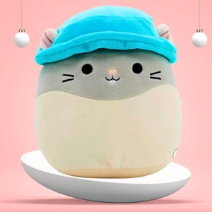 May include: A plush toy shaped like a hamster wearing a blue hat. The toy has a gray face with black eyes and a pink nose. The toy is sitting on a white platform.