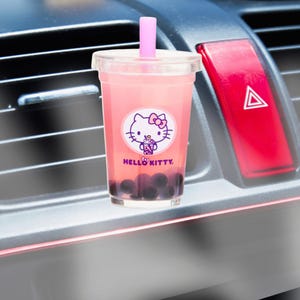 Pode incluir: Um copo de plástico transparente cheio de bubble tea rosa, com um desenho da Hello Kitty. O copo tem um canudo rosa e contém pérolas de tapioca escuras. O logotipo da Hello Kitty está em um círculo branco com as palavras "Hello Kitty" abaixo.