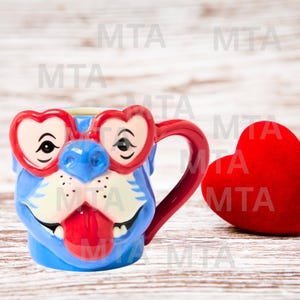 May include: A blue ceramic mug shaped like a dog's face, wearing red heart-shaped glasses. The mug has a red handle and tongue, with a red heart-shaped plush next to it. The mug has the text "MTA" repeated on the background.