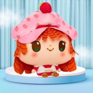 May include: A plush doll with a strawberry theme. The doll has a pink hat with strawberry patterns and a red strawberry on top. It has orange hair, large brown eyes, and a smiling face. The doll is wearing a white and red outfit.