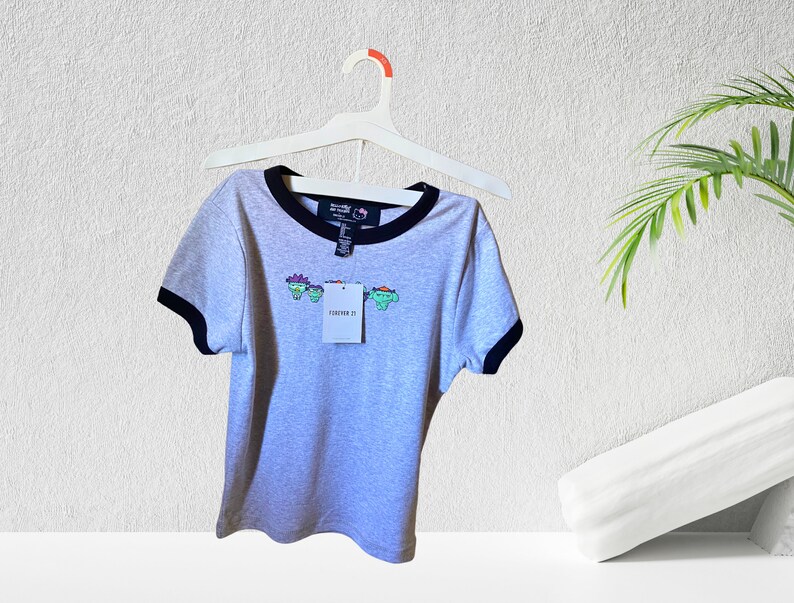 May include: Gray short-sleeved t-shirt with a black and white ringer neckline. The shirt features a graphic of four cartoon characters with the text "Forever 21" on the front.