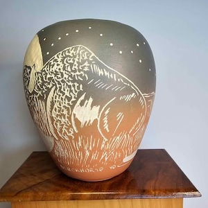 May include: A large, rounded ceramic vase with a hand-painted design. The vase features a bison illustration in white against a gradient background of gray and orange. The top has white dots, and the bottom has the text "BLACKHORSE 10".