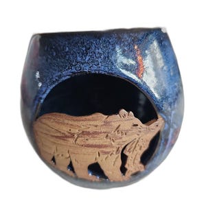 May include: A round ceramic candle holder with a deep blue, mottled glaze finish. The front features a large circular cutout containing a raised, unglazed brown clay relief of a bear walking through a forest setting with trees.