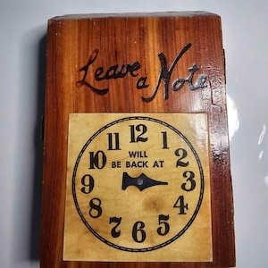 May include: A rectangular wooden wall sign featuring the text Leave a Note at the top and a clock face below with the text WILL BE BACK AT. The clock face has black numbers from 1 to 12 and a single black hand pointing toward the number 3.