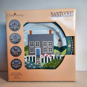 Op de afbeelding: Een set van vier salade/dessert borden met een Nantucket ontwerp, gepresenteerd in een kartonnen doos. De borden tonen een grijs huis met een blauwe deur, een windmolen en bloemmotieven. De doos is beige en draagt de logo's "Claire Murray" en "Nantucket".