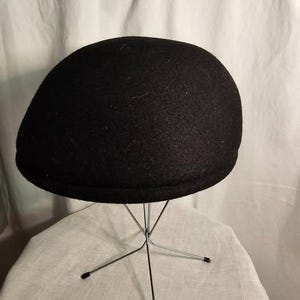 Peut inclure: Casquette de style gavroche noire en laine. Le chapeau est présenté sur un support métallique avec une base en forme d'étoile. La casquette a une couronne arrondie et un petit bord rigide. Le tissu est de couleur foncée unie.