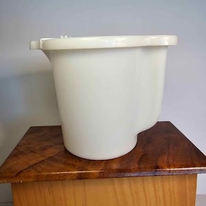 Può includere: Un secchio di plastica bianca con manico e design curvo. Il secchio è appoggiato su una superficie di legno.