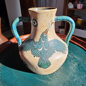 Op de afbeelding: Een keramische vaas met een beige achtergrond en twee blauwe handvatten. De vaas heeft een blauwgroene vogel met uitgespreide vleugels. De vogel heeft witte accenten en een open bek. De handvatten zijn effen blauw.