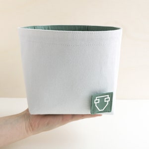A hand of caucasian woman holding a white fabric box with a pastel green inside and a square label with a white clipart of a diaper.