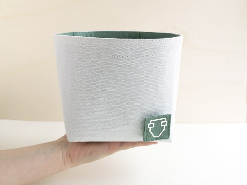 A hand of caucasian woman holding a white fabric box with a pastel green inside and a square label with a white clipart of a diaper.