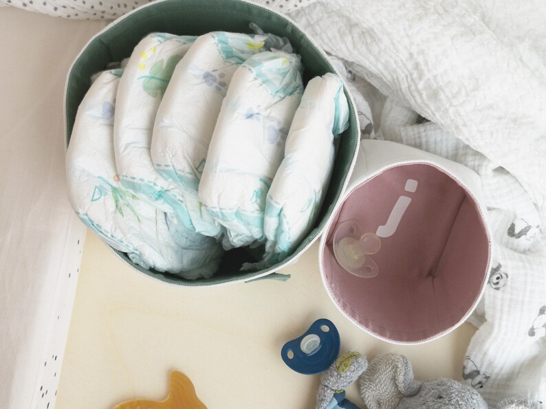 Lifestyle photo of two white fabric boxes from above. The bigger one on the left has pastel green inside - diapers are inside. The smaller one on the right has pastel rose inside and white letter j and a pacifier is inside.