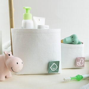 Lifestyle photo of two white fabric boxes. The bigger one on the left with green label with white clipart of baby face - different toiletries are inside. The smaller one on the right with rose label with white clipart of duck - bath toy inside.