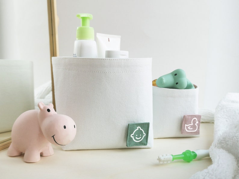 Lifestyle photo of two white fabric boxes. The bigger one on the left with green label with white clipart of baby face - different toiletries are inside. The smaller one on the right with rose label with white clipart of duck - bath toy inside.