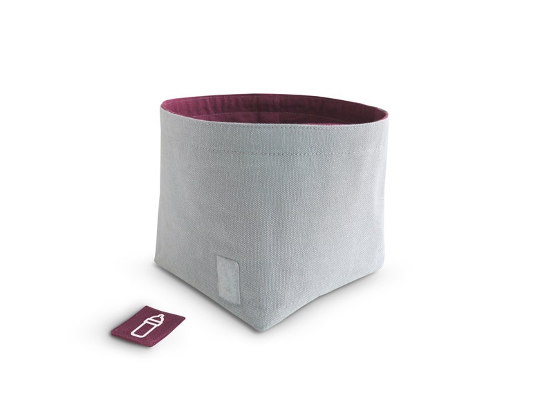 Front view of grey fabric storage bin with mulberry pink inside and grey rectangle hook-and-loop fastener on the front side. The mulberry pink square label with white clipart of milk bottle lays infront of the basket.