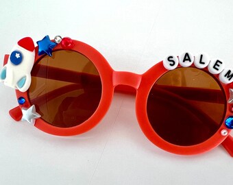 May include: Red sunglasses with brown lenses, featuring a white rocket, stars, and the word "SALEM" in white beads. The sunglasses have a decorative design.