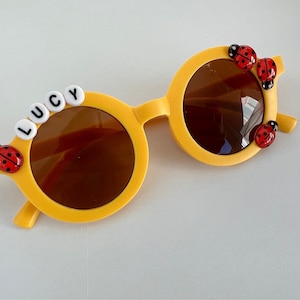 May include: Yellow sunglasses with brown lenses, adorned with red ladybug details. The name "LUCY" is spelt out in white letter beads across the top. A cheerful and playful accessory.