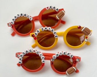 May include: Three pairs of round sunglasses in orange and yellow. Each pair has a beaded message: "FOOTBALL", "GO CHIEFS", and "KANSAS CITY CHIEFS". Each pair has a football charm and decorative rhinestones.