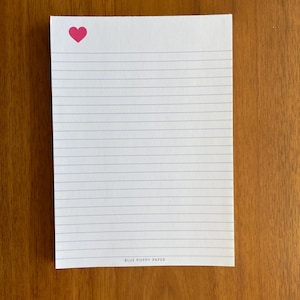 May include: White lined notepad with a pink heart in the upper left corner. The notepad has a simple design with horizontal blue lines for writing. The words "BLUE POPPY PAPER" are printed at the bottom of the notepad.