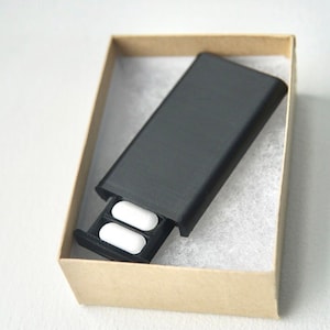 Puede incluir: Un pastillero negro con dos pastillas blancas en el cajón abierto, presentado dentro de una pequeña caja de cartón abierta. La caja está forrada con material blanco y esponjoso. El estuche es rectangular y tiene un cajón deslizante.