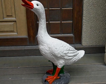 May include: A white goose statue with a bright orange beak and orange webbed feet. The goose is depicted in a standing position with its head tilted upward and its beak open. The figure is mounted on a small, dark gray, textured base.