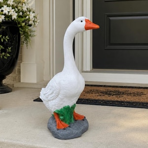 May include: A white goose garden statue with an orange beak and orange webbed feet. The goose is standing on a gray rock base with a small patch of green grass at its feet. The figure is sculpted with detailed feather textures on its body.