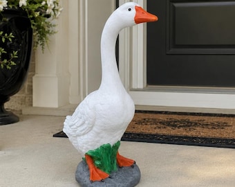 May include: A white goose garden statue with an orange beak and orange webbed feet. The goose is standing on a gray rock base with a small patch of green grass at its feet. The figure is sculpted with detailed feather textures on its body.