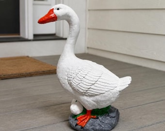 May include: A white goose figurine with a bright orange beak and orange webbed feet. The goose stands on a textured gray rock base with a small white egg nestled near its feet. The sculpture features detailed feather textures across its body.